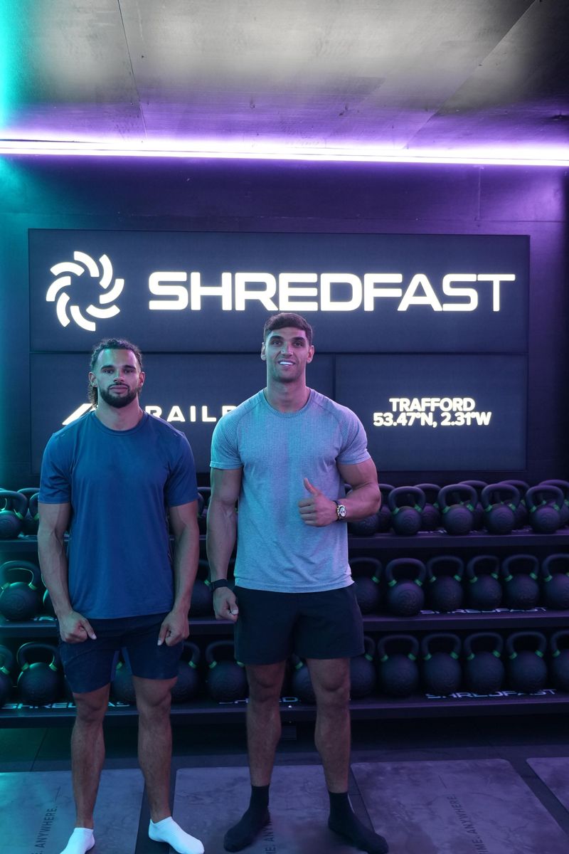 Tyler and Reuben, owners of ShredFast Trafford, in front of the illuminated ShredFast Trafford sign and kettlebell racks